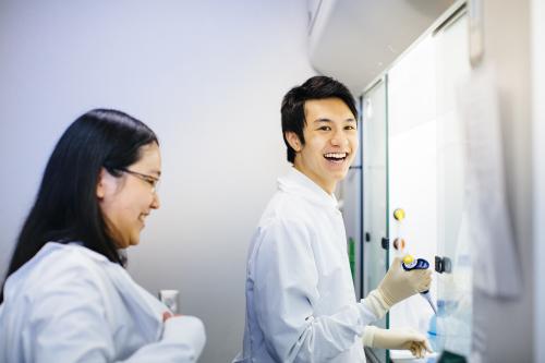 A UCLA researcher stands smiling with a pipet while another looks on.