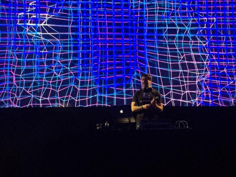 Dr. Tony Aldave wears a black T-shirt and holds a microphone on a vibrant DJ stage.