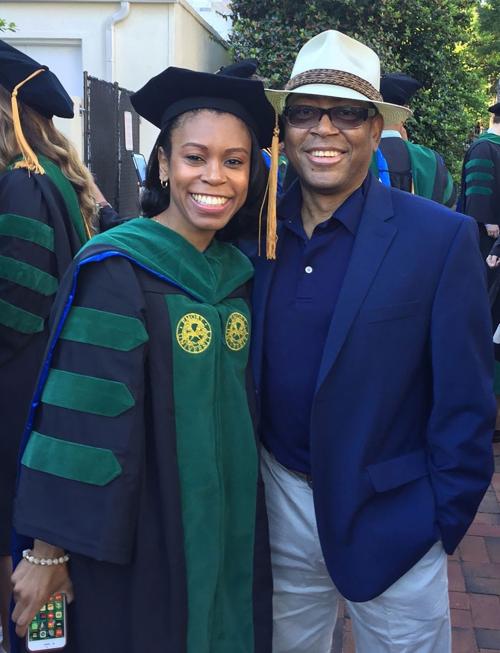 A Patrice in academic graduation regalia with green and black doctoral robes and mortarboard poses with Mann in a blue suit and panama hat at what appears to be a graduation ceremony outdoors.