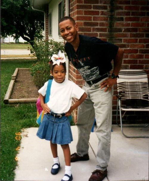 Mann in casual clothing (dark shirt and light pants) poses with his daughter, Patrice, wearing a white shirt and blue skirt with a hair bow, standing on a patio or walkway in front of a brick building.