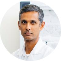 Headshot of Ranmal Samarasinghe, a man with short dark hair wearing a white lab coat, with a bright laboratory environment in the background.
