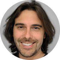 Headshot of Daniel Aharoni, a man with medium-length dark brown hair and a beard, smiling warmly against a gray background.