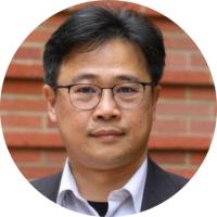 Headshot of Eric Chiou with black hair and wire-rimmed glasses wearing a white shirt and dark suit jacket in front of a brick wall.