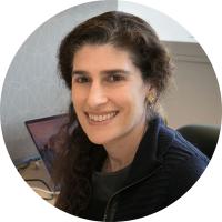 Headshot of Hilary Coller, a woman with long curly dark brown hair wearing gold earrings and a dark jacket, smiling in an office setting with a laptop visible in the background.