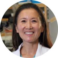 Professional headshot of a woman in a white lab coat over blue scrubs, smiling in a laboratory setting