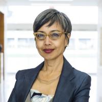 Professional headshot of Ninez A. Ponce, Ph.D., M.P.P., wearing a dark blazer and yellow-framed glasses, smiling against a bright office background