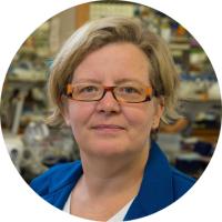 Headshot of Kathrin Plath with short blonde hair and rectangular orange glasses wearing a blue lab coat in a laboratory setting.
