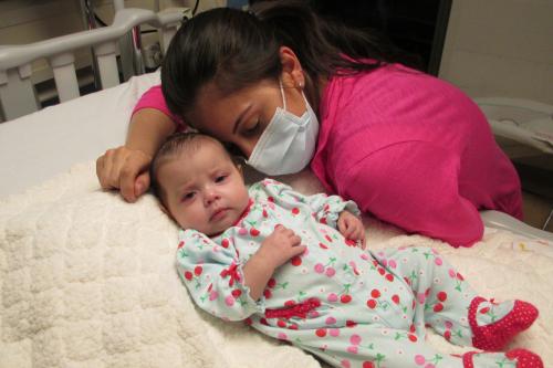 A baby lies in a hospital bed with her mother in a face mask