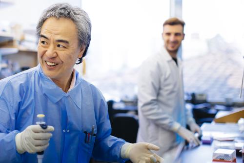 Two scientists stand smiling, working at a lab bench. One holds a pipet.