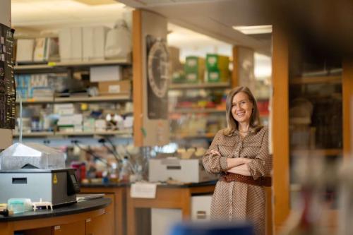 Hanna Mikkola in a brown patterned coat stands with crossed arms, smiling in a laboratory filled with scientific equipment and supplies.