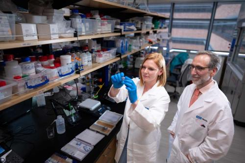 Two UCLA scientists are photographed in the lab together. Katie Campbell, PhD, wears a labcoat and blue gloves, and Antoni Ribas, MD, PhD. also wears a white labcoat and stands with his hands in his pockets.