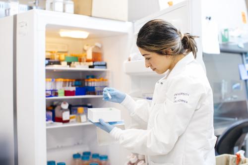 A scientist looks at a vial of liquid in front of a refrigerator in a lab