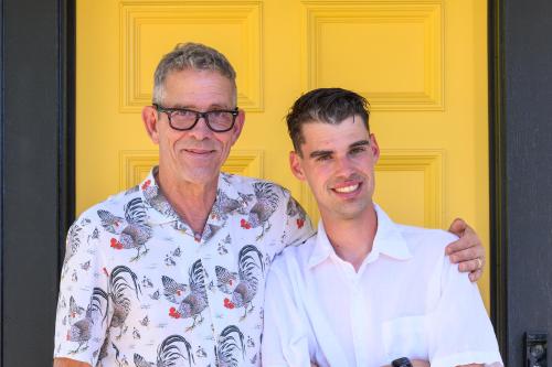 Two men standing together and smiling in front of a bright yellow door - Joe Henry wearing glasses and a patterned shirt with his arm around his son, Franklin, in a white shirt.