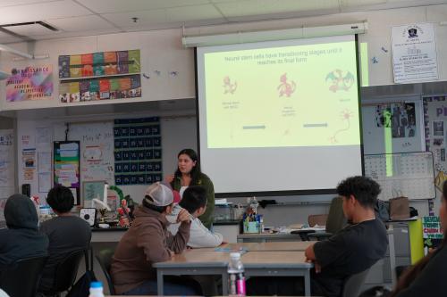Jessenya Mil presents a slide about neural stem cell development to students seated in a classroom, with the slide using Pokémon-style evolution graphics to illustrate cell stages.