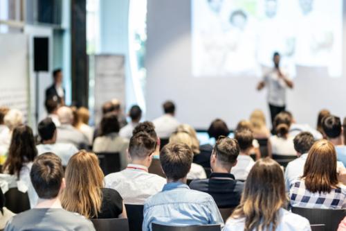 A large audience listening to a person on stage giving a presentation. 