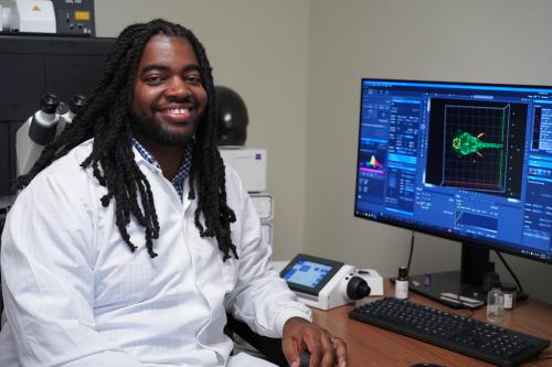 Headshot of Dr. D'Juan Farmer wearing a white lab coat sitting next to a computer monitor with zebrafish on the screen