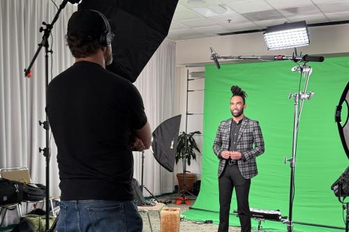 Behind-the-scenes photo of an elevator speech video shoot: a trainee in a plaid blazer faces the camera in front of a green screen, flanked by studio lighting, as a videographer looks on.