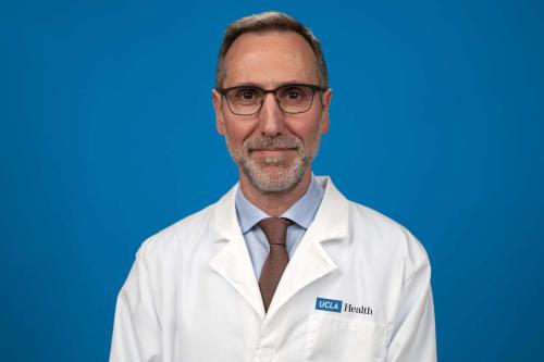 UCLA's Dr. Antoni Ribas smiles for a photo. He wears a whitecoat and glasses and poses against a blue background.