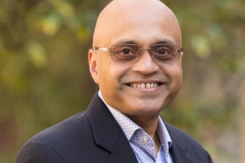 UCLA's Dr. Arjun Deb smiles for a headshot outdoors. He wears a pair of glasses and a light blue shirt and suit jacket.