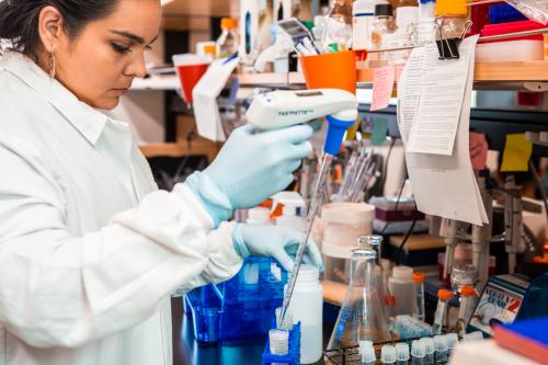 A female scientist stands pipetting at a lab bench cluttered with equipment. 