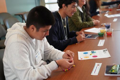 a male high school student is making a bracelet seated next to another male high school student