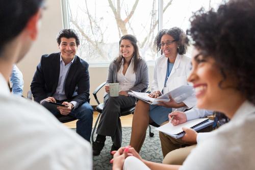 A group of doctors sit in a circle indoors, smiling and speaking to each other. They are dressed either in white coats or in business attire, and some hold notebooks, binders, or tablets in their hands. 
