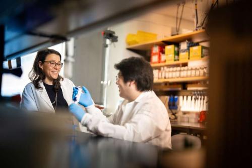 Dr. Cristina Puig-Saus is pictured wearing a white lab coat while standing in a laboratory and speaking with another scientist. Both individuals are smiling. 