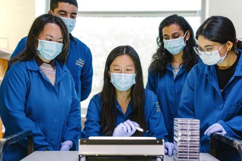 Dr. Melody Li, centered, gestures to a benchtop instrument while four masked researchers in blue lab coats watch closely. A stack of clear culture plates sits on the lab bench beside them.