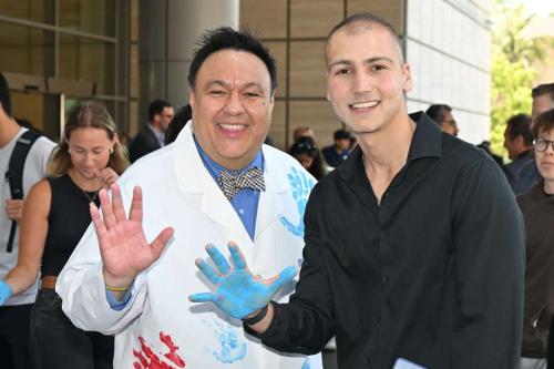 a photo with pediatric cancer researcher Dr. Steve Jonas wearing a white coat on the left posing with cancer survivor Sina Sinbari on the right wearing a black collared long sleeve shirt at UCLA Mattel Children's Hospital