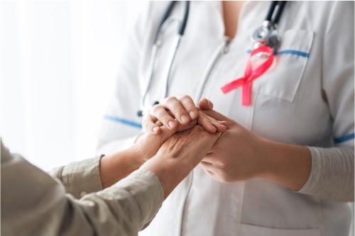A patient, left, and medical care provider holding hands; provider has a pink breast cancer ribbon on her lapel