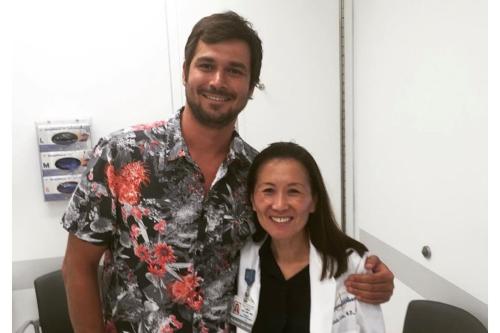 Attorney and brain cancer survivor Jamil Newirth stands smiling beside Dr. Linda Liau, chair of neurosurgery at the David Geffen School of Medicine at UCLA and director of the UCLA Brain Tumor Program, in a medical office setting. Newirth wears a colorful floral shirt while Dr. Liau wears a white medical coat.