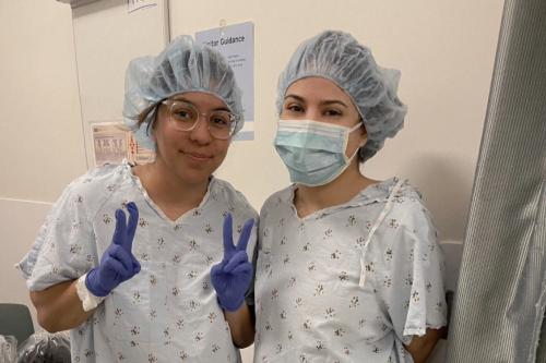 Two sisters in hospital gowns and hair coverings prepare for surgery.