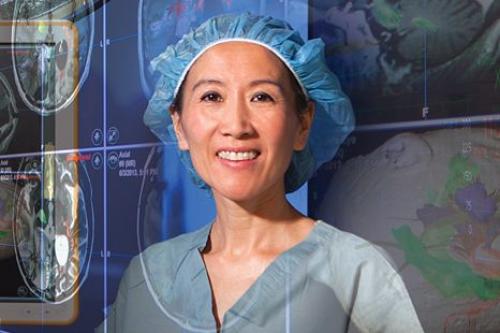 Dr. Linda Liau in surgical scrubs and cap standing in an operating room with brain scan images and surgical lighting visible in the background.