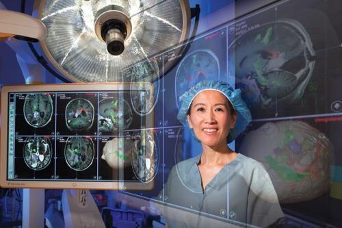 Dr. Linda Liau in surgical scrubs and cap standing in an operating room with brain scan images and surgical lighting visible in the background.