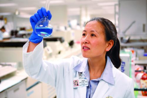 Dr. Linda Liau in a white lab coat and blue gloves examining a petri dish in a laboratory.