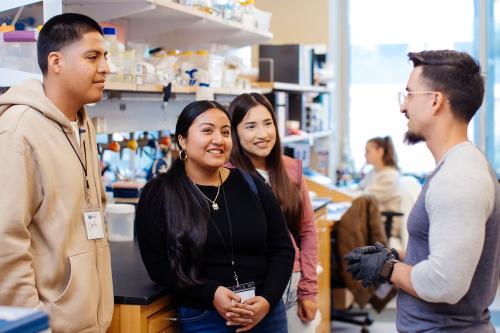 three high school students are standing in a research lab facing a male ucla researcher
