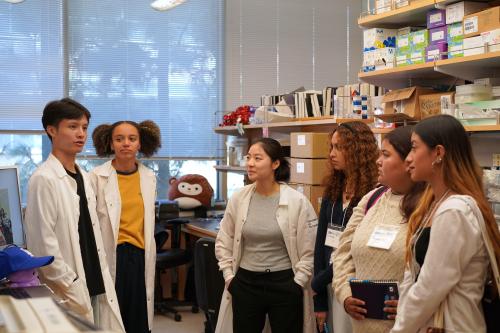 three scientists wearing white lab coats stand in between a bay talking to three female high school students