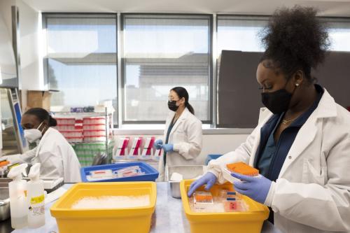 Three masked researchers in labcoats work at various stations in a UCLA lab. 