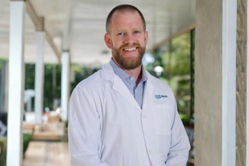UCLA researcher Vondriska, Thomas smiles for a photograph. He wears a white UCLA labcoat and stands indoors with a green landscape appearing behind him.