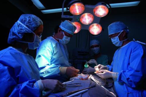A group of surgeons gather around a patient in an operating room. The lights are dimmed and blue-tinged and overhead light blares above.