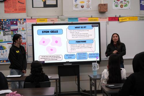 two female researchers stand in front of a high school classroom with a TV monitor in between them projecting a slide on stem cells