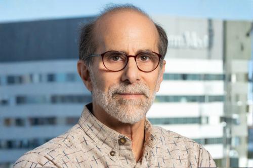 Headshot of Kenneth Zaret in a tan button up and glasses against a cityscape