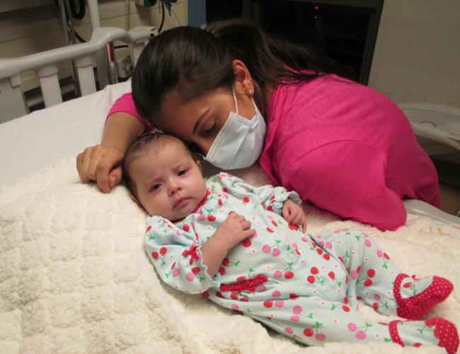 A baby lies in a hospital bed with her mother in a face mask