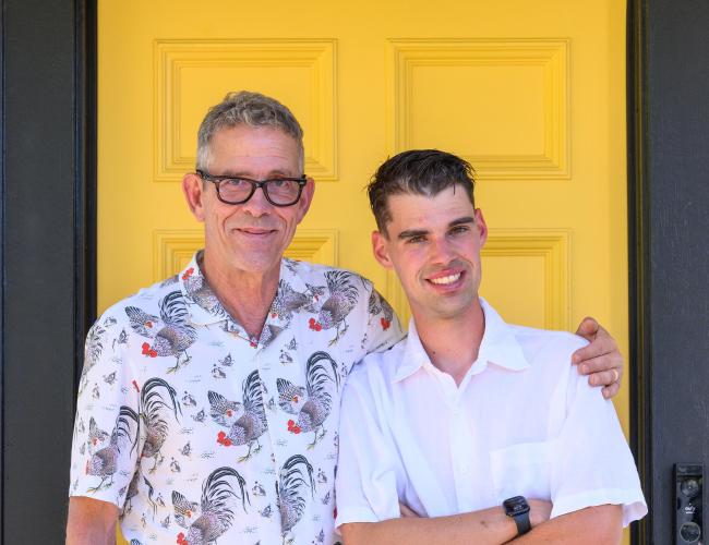 Two men standing together and smiling in front of a bright yellow door - Joe Henry wearing glasses and a patterned shirt with his arm around his son, Franklin, in a white shirt.
