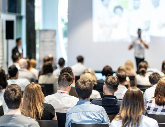 A large audience listening to a person on stage giving a presentation. 