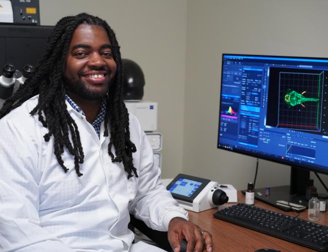 Headshot of Dr. D'Juan Farmer wearing a white lab coat sitting next to a computer monitor with zebrafish on the screen