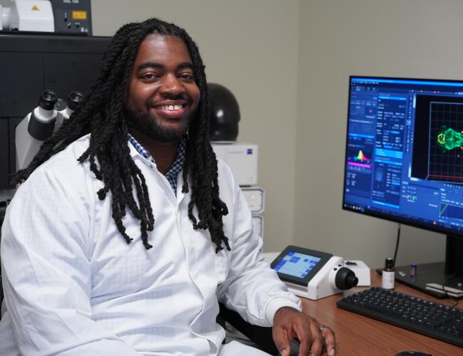 Headshot of Dr. D'Juan Farmer wearing a white lab coat sitting next to a computer monitor with zebrafish on the screen