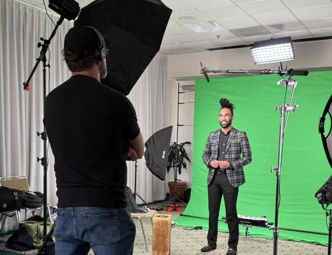 Behind-the-scenes photo of an elevator speech video shoot: a trainee in a plaid blazer faces the camera in front of a green screen, flanked by studio lighting, as a videographer looks on.
