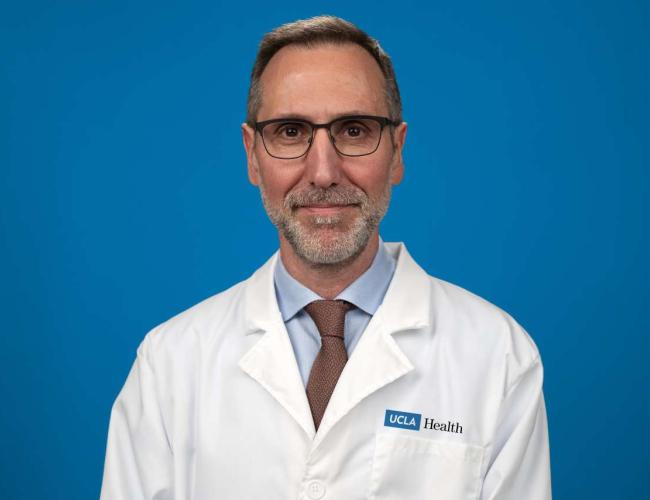 UCLA's Dr. Antoni Ribas smiles for a photo. He wears a whitecoat and glasses and poses against a blue background.