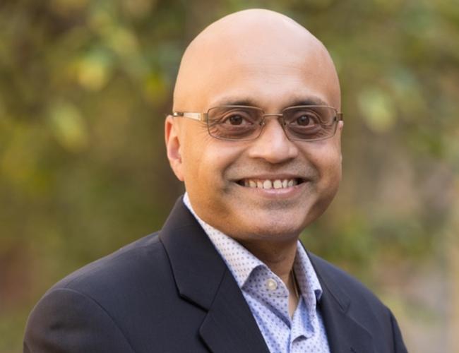 UCLA's Dr. Arjun Deb smiles for a headshot outdoors. He wears a pair of glasses and a light blue shirt and suit jacket.